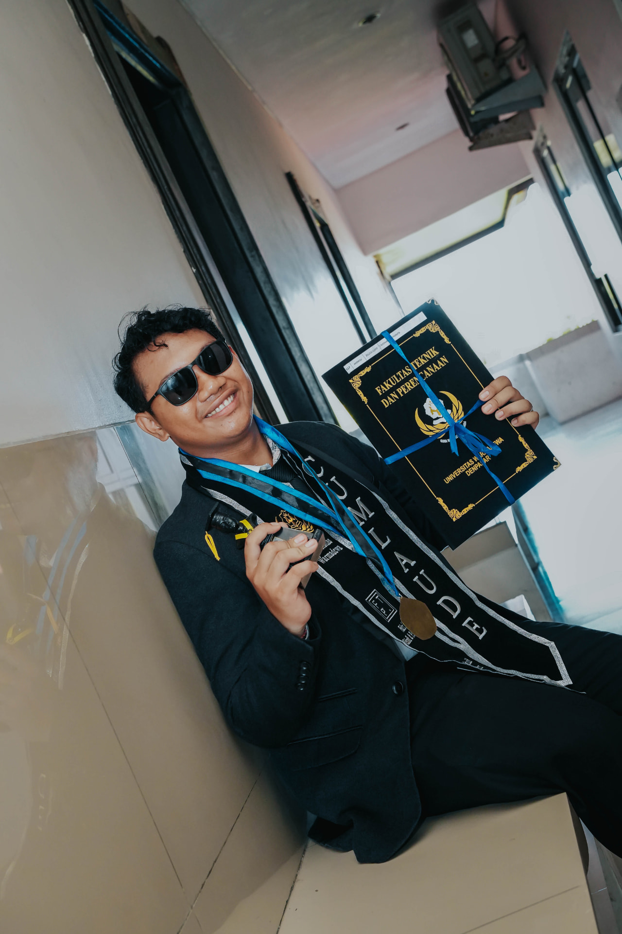 Bali graduation photography — proud graduate in gown holding his cum laude diploma and medal