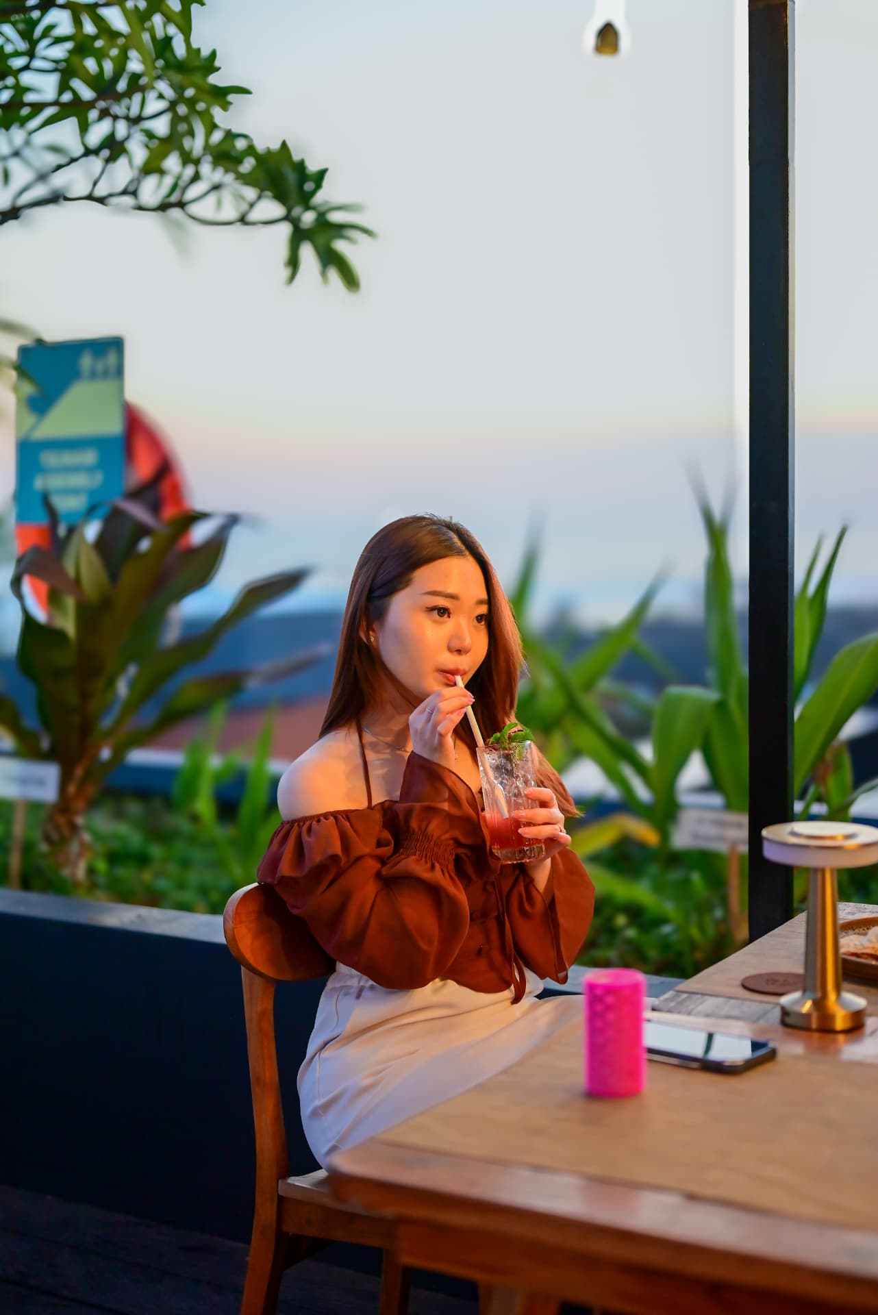 Bali lifestyle portrait — woman enjoying a drink at a rooftop café in golden evening light